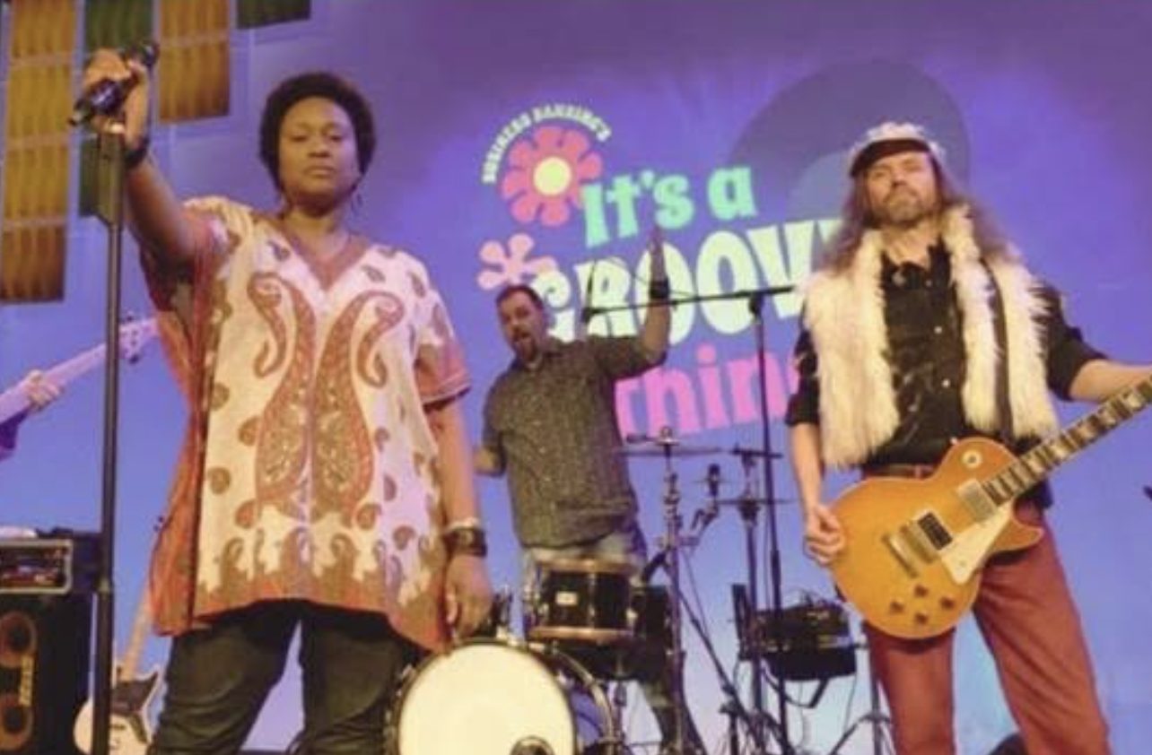 Three musicians performing on stage with a colorful retro-style backdrop