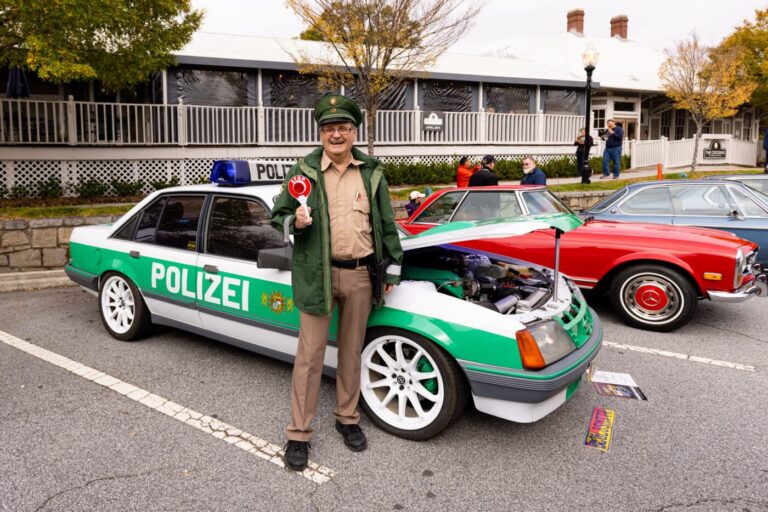 A man in a green uniform stands in front of a green-and-white police-themed car at a car show, with classic cars parked beside him.