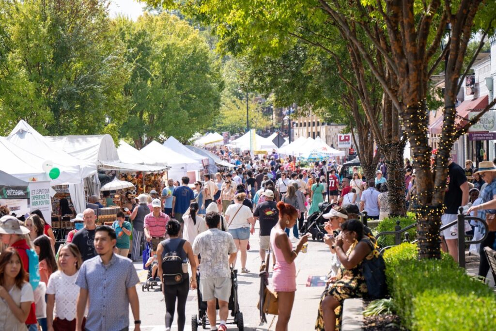 Crowds walk past rows of white vendor tents along a tree-lined downtown street.