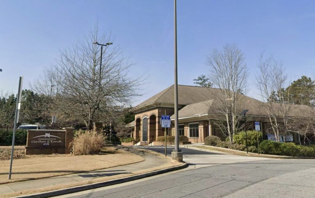 A one-story synagogue building beside a driveway and parking signs, with a sign at the curb.