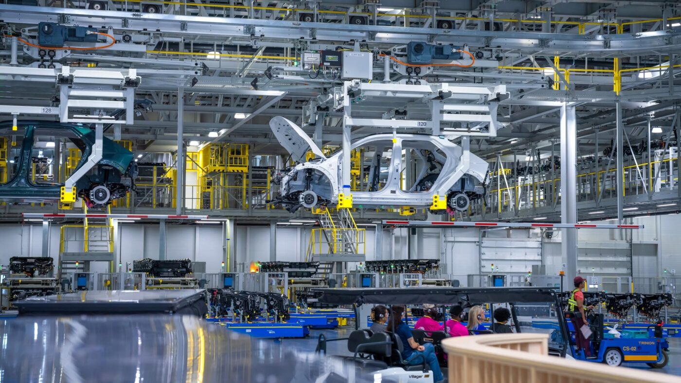 Línea de ensamblaje dentro de Hyundai Motor Group Metaplant America con carrocerías de vehículos suspendidas en el techo y trabajadores abajo.