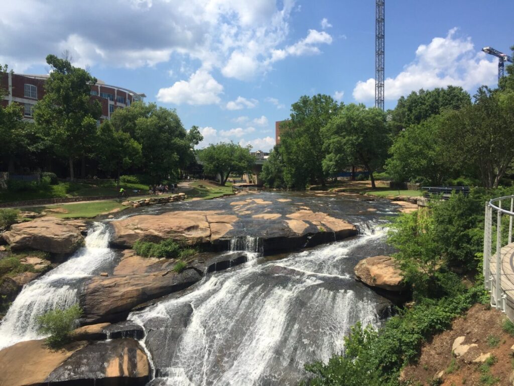 A broad waterfall spills over flat rock ledges in a park with trees and a city skyline nearby.