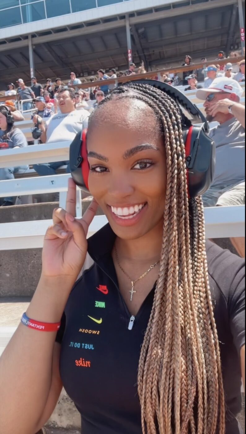 A smiling woman with long braided hair wears large earmuffs and holds up a peace sign at an outdoor event.