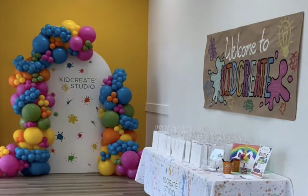 Colorful balloon arch frames a Kidcreate Studio doorway beside a welcome sign and a table set with favors.
