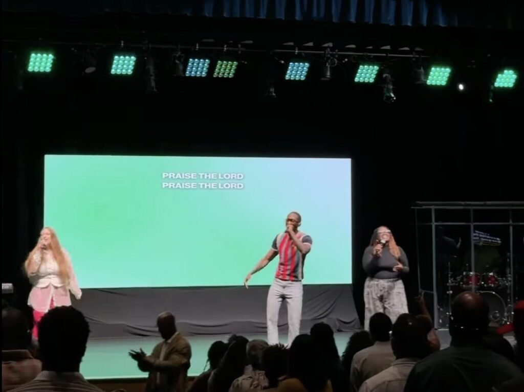 Three worship leaders on a church stage in front of a large screen and seated audience