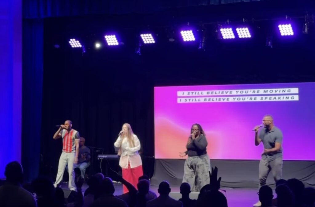 Four singers on a church stage under purple lighting with audience silhouettes in front