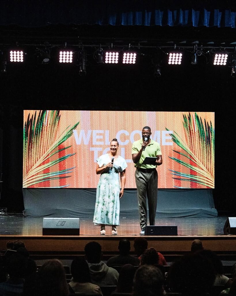 Two people holding microphones on a church stage in front of a welcome screen and seated audience