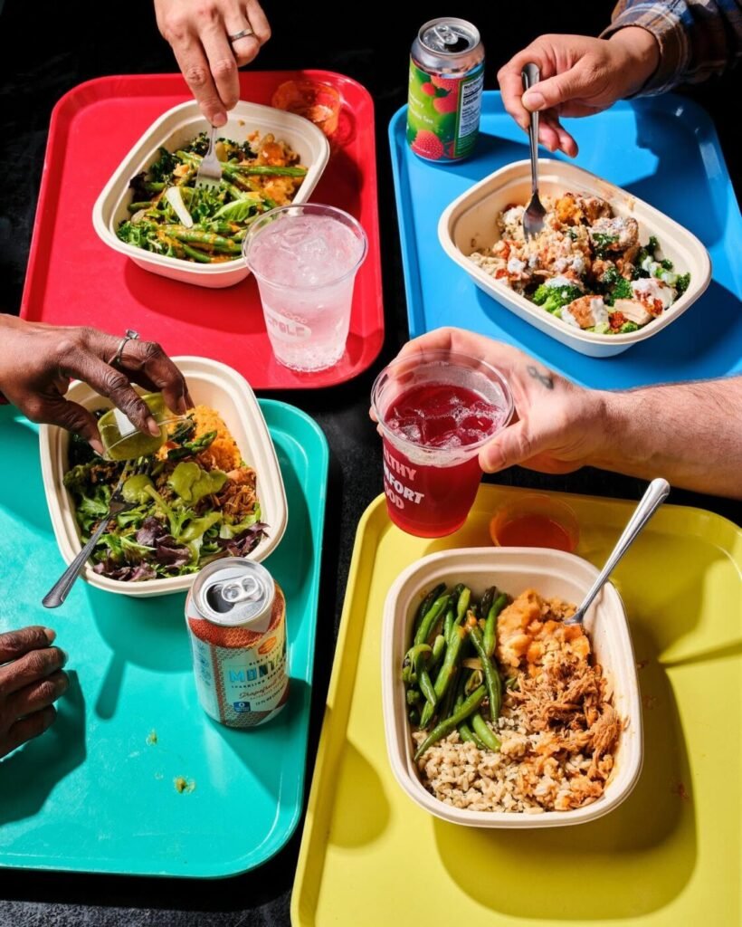 Four people reach toward colorful meal trays holding takeout bowls and drinks