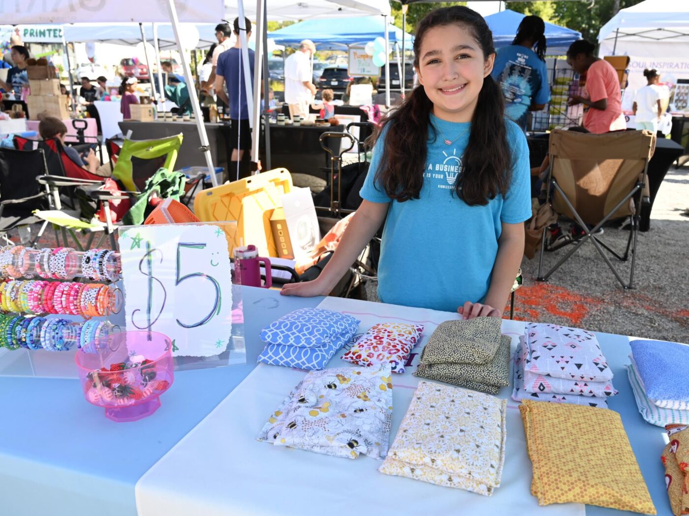 A girl stands behind a table of handmade items for sale, including folded fabric goods and bracelets with a handwritten five-dollar sign.