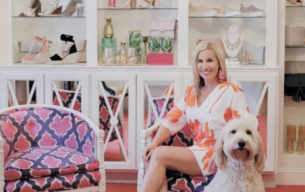 Monkee’s boutiques lean into a full-service shopping experience with clothing, shoes and accessories in one place. A woman in a floral outfit sits in a boutique showroom beside a white dog, with shelves of shoes and accessories behind them.