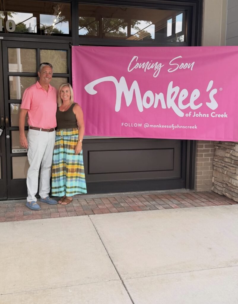 A man and woman stand beside a bright pink Monkee’s of Johns Creek coming soon banner outside a storefront.