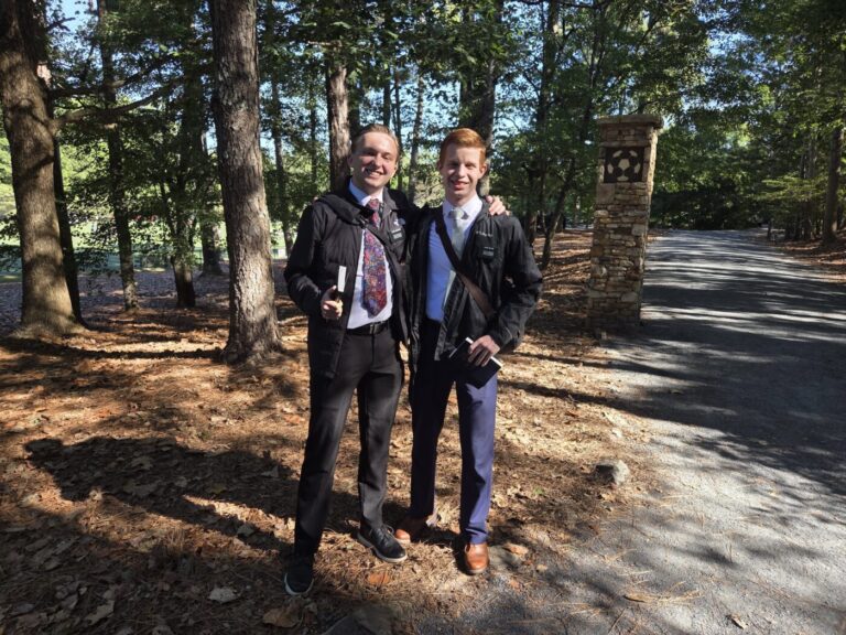 Two young men in dark jackets and ties stand together on a wooded park path, each holding a small book.
