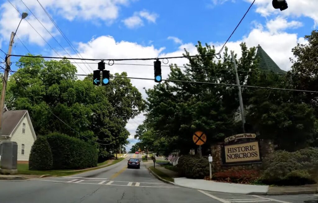A car drives along a tree-lined street past a Historic Norcross sign and overhead traffic lights.