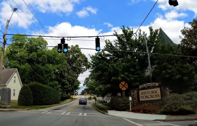 A car drives along a tree-lined street past a Historic Norcross sign and overhead traffic lights.