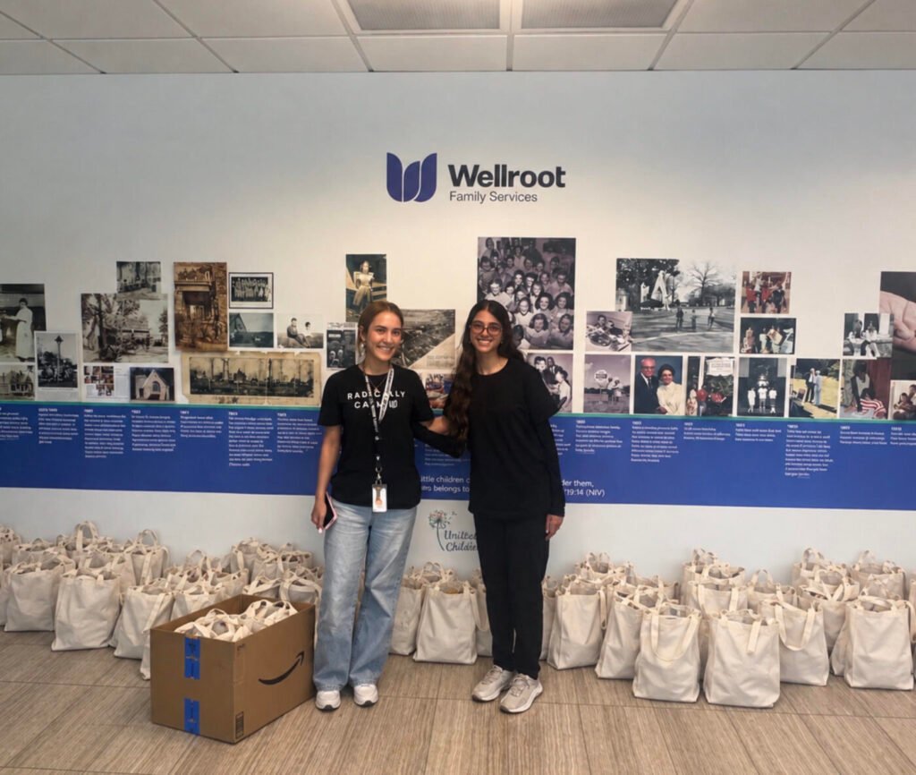 Two women stand in front of a Wellroot Family Services wall display, with many tote bags lined up behind them.