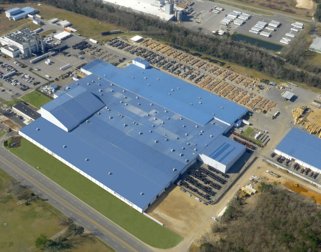A large blue-roofed industrial building surrounded by parking lots, storage yards and trucks, seen from above