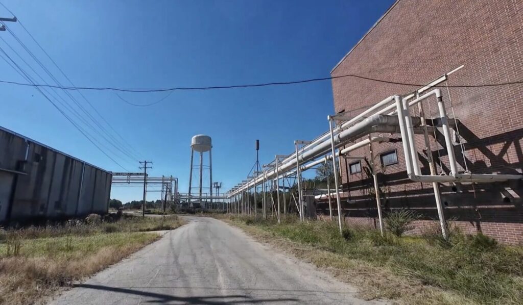 An empty road runs between industrial buildings and elevated pipes near a water tower under a clear blue sky.