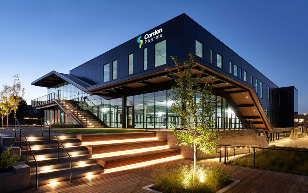 A modern CordenPharma building with glass walls and illuminated steps at dusk