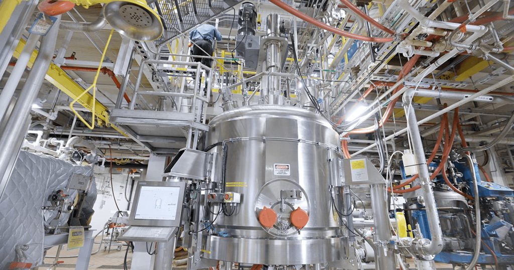 A stainless steel pharmaceutical manufacturing tank surrounded by pipes, platforms and overhead equipment