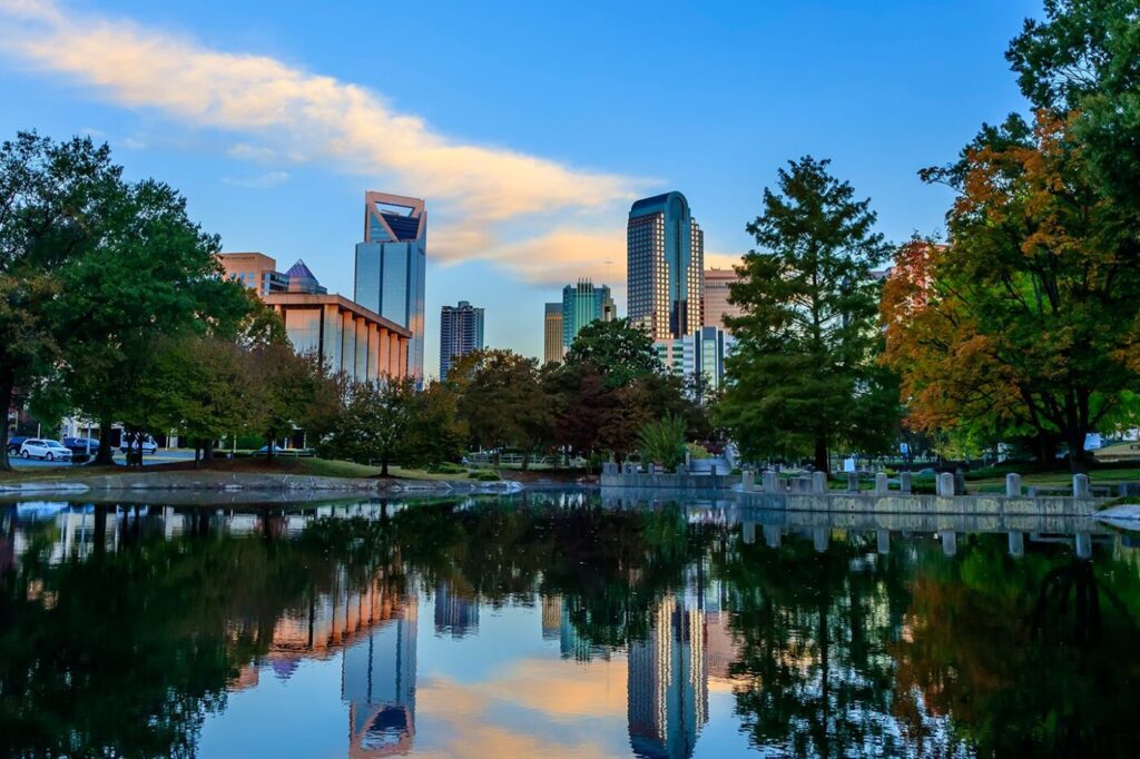 Charlotte skyline reflected in a calm pond at sunset, framed by trees on both sides.