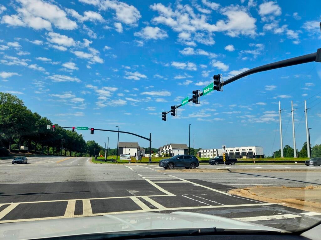 Wide intersection with traffic lights, crosswalk markings and several cars under a blue sky.