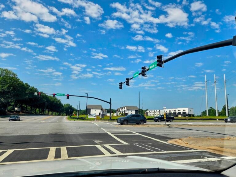 Wide intersection with traffic lights, crosswalk markings and several cars under a blue sky.