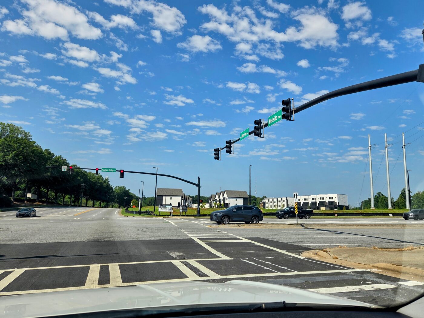 Wide intersection with traffic lights, crosswalk markings and several cars under a blue sky.