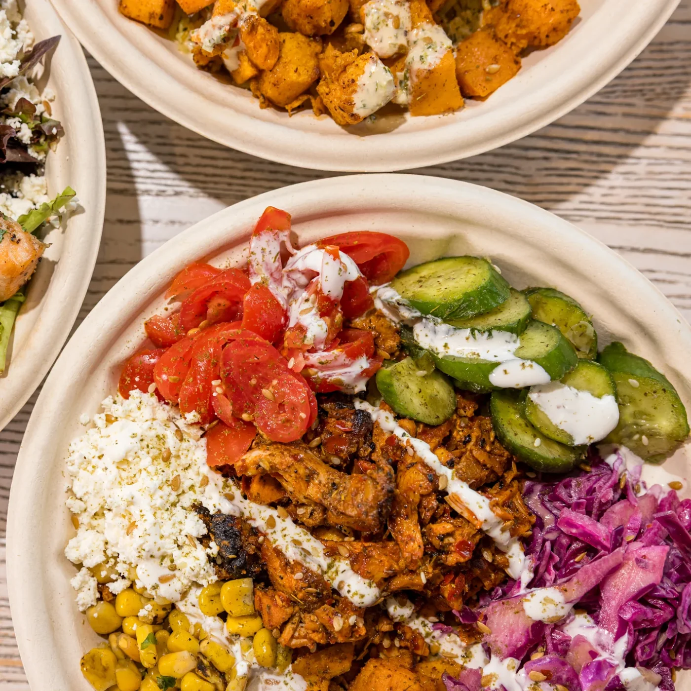A bowl of grains topped with chicken, cucumber, tomatoes, cabbage, corn and white sauce