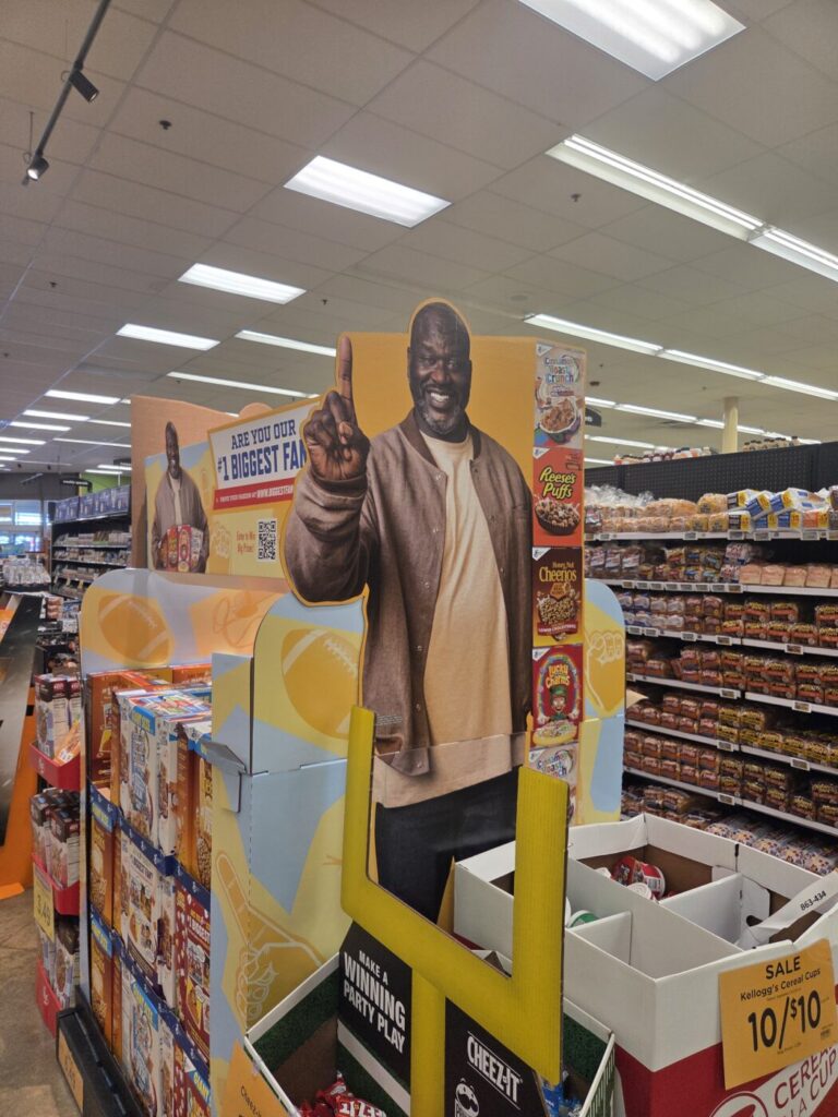 A cardboard cutout of Shaquille O'Neal stands in a grocery store cereal aisle beside stacked boxes of cereal.