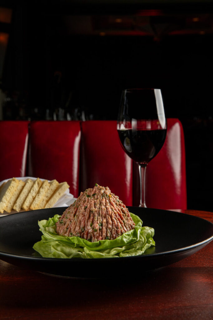 A plate of steak tartare garnished with lettuce sits beside a glass of red wine and sliced bread in a dim restaurant setting.
