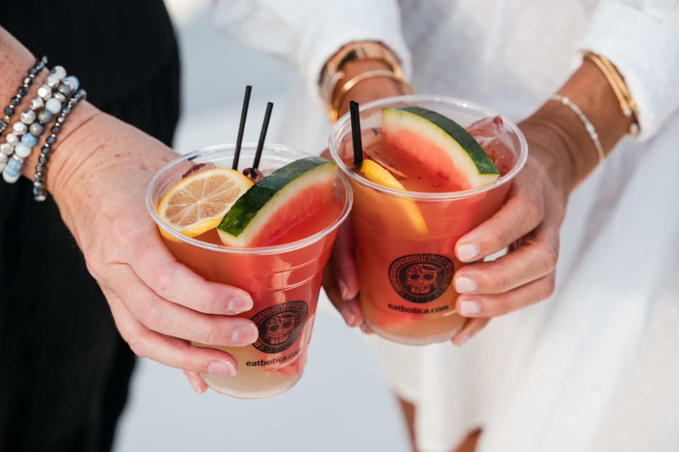 Two people hold plastic cups of pink cocktails garnished with watermelon slices, lemon, and black straws.
