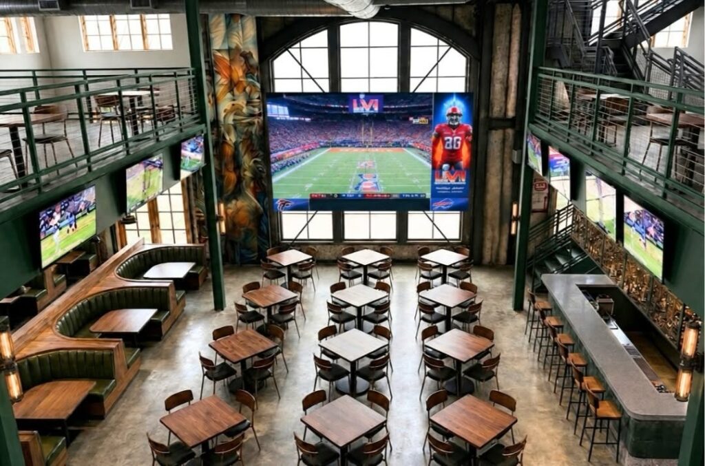 High-ceiling restaurant interior with a large central television surrounded by smaller screens, tables and a bar