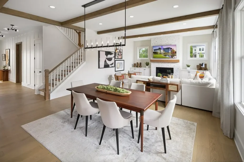 A staged dining area and living room with white sofas, wood beams and a staircase leading upstairs.