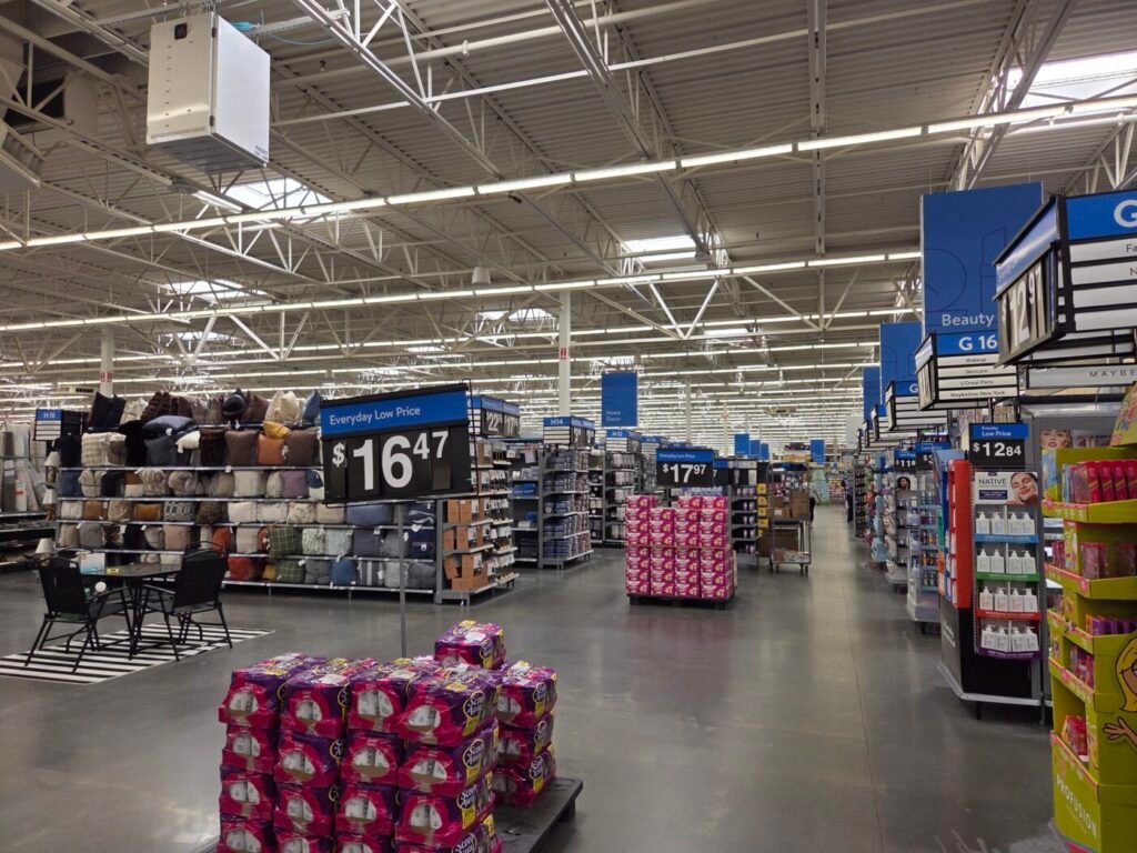 A wide aisle inside a Walmart store with overhead signs, stacked merchandise and bright warehouse-style lighting.