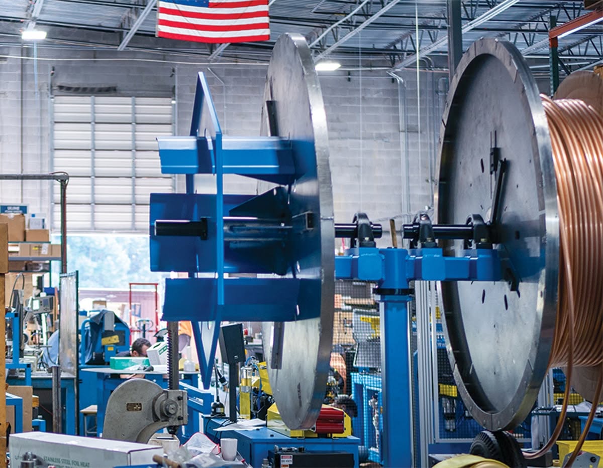 Blue industrial machinery inside a factory, with large metal reels and an American flag hanging overhead.