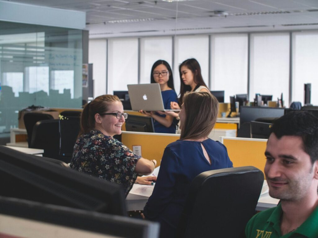 People working at desks in an office while two women stand nearby with a laptop.