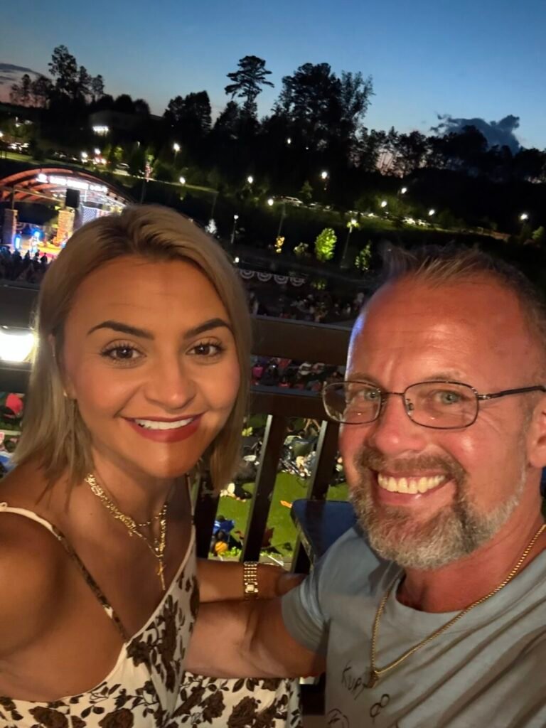Smiling man and woman posing for a selfie with a lit amphitheater and crowd in the background at dusk