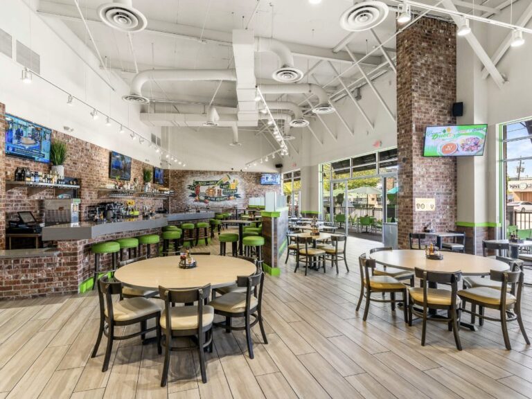 Bright dining room with round tables, green bar stools and a long bar under high white ceilings