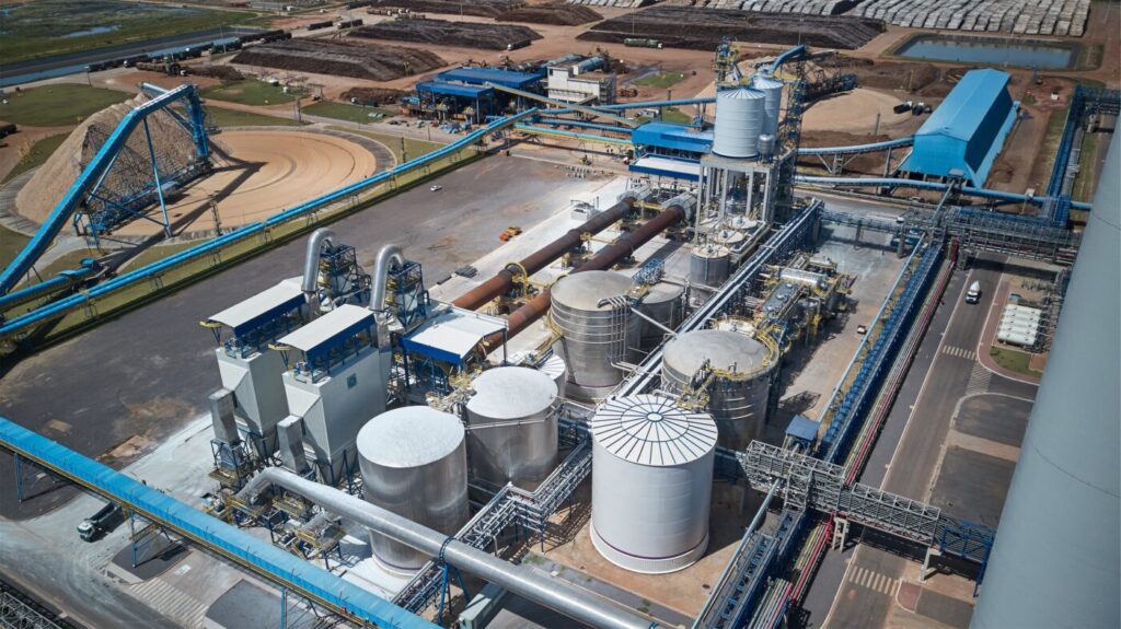 Aerial view of an industrial plant with large cylindrical tanks, piping, conveyors and blue-roofed structures.