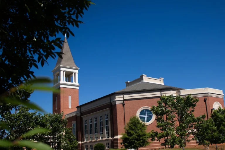 Red-brick church building with a tall steeple framed by trees under a blue sky.