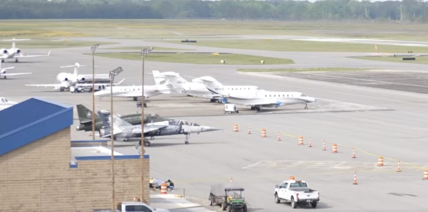 Several aircraft parked on an airport apron beside taxiways and a low blue-roofed building.