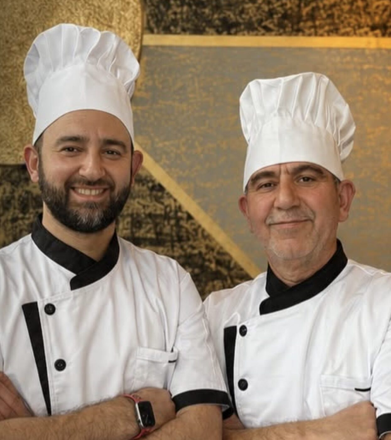 Two chefs in white jackets and tall white hats stand side by side and smile.