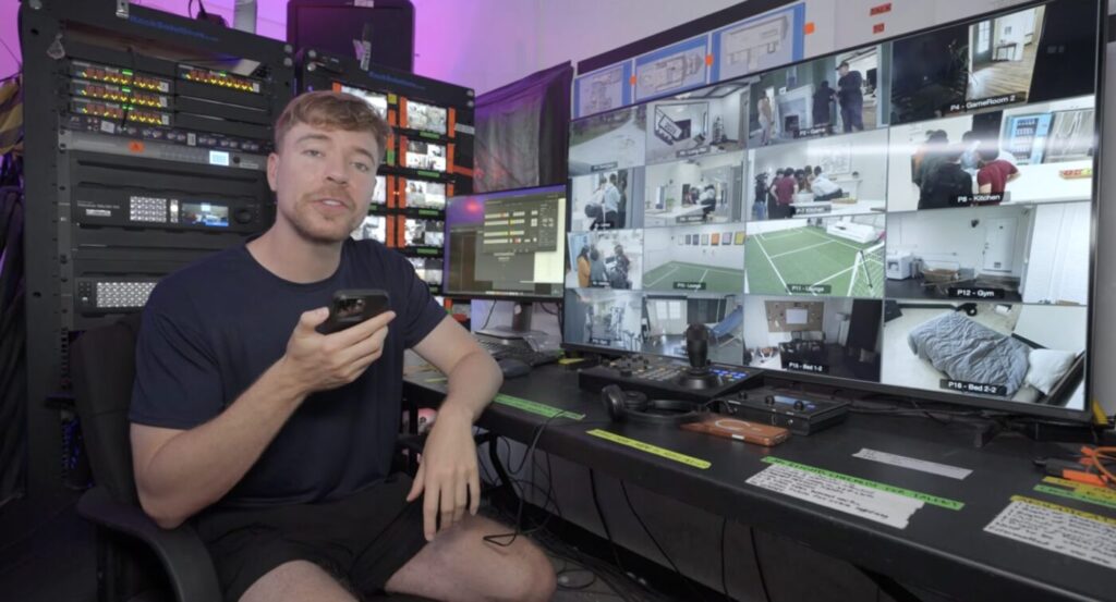 Jimmy Donaldson sits in front of a wall of security monitors and equipment while holding a phone.