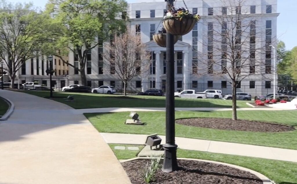 Walkways and landscaping outside a government building with columns and parked cars along the street.
