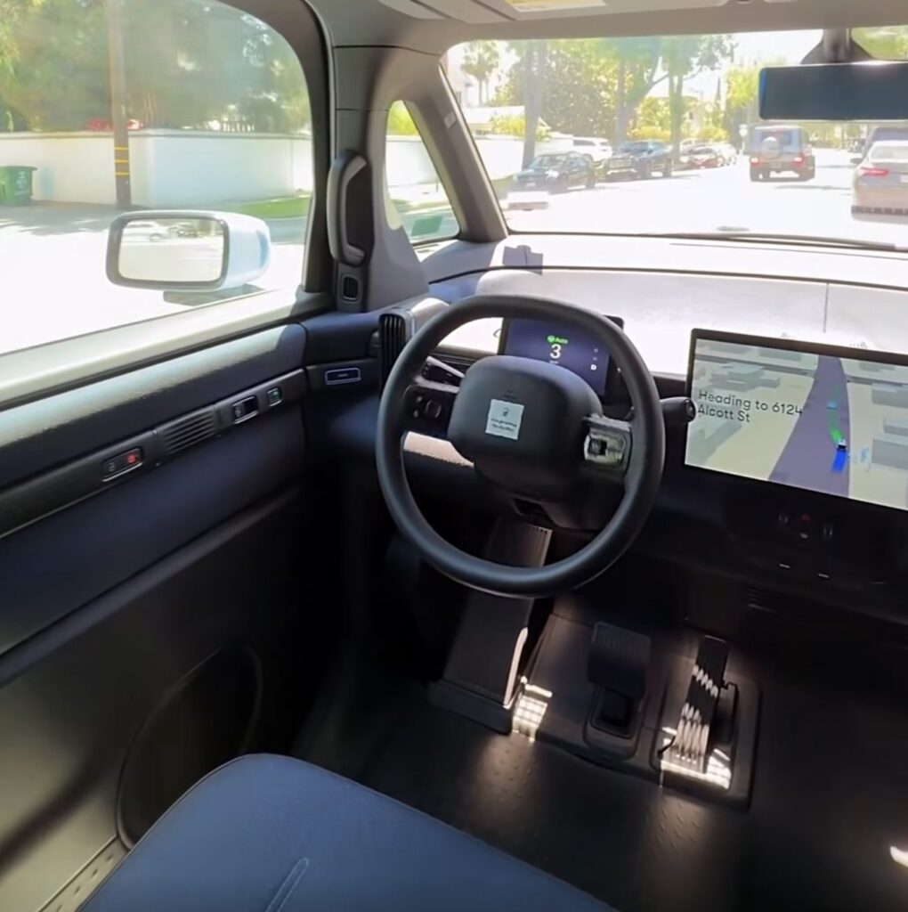 Interior view of a Waymo vehicle with a steering wheel, front seat, and passenger screen showing a route map.