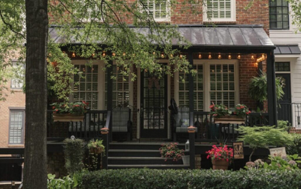 A brick house with a covered front porch and potted flowers sits behind leafy trees.