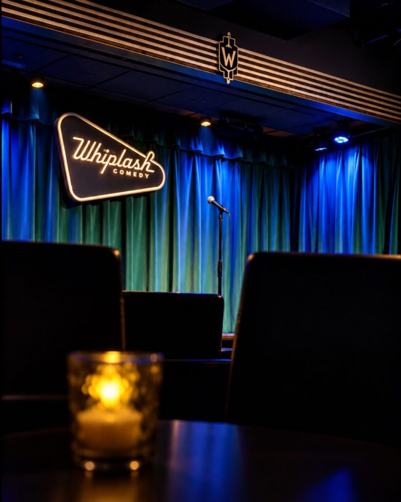 A microphone stands alone on a small stage in front of blue curtains, with a Whiplash Comedy sign glowing nearby and empty seats in the foreground.