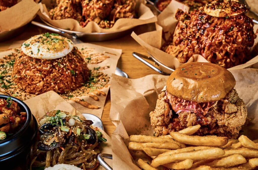 Two plated fried chicken dishes, a chicken sandwich and fries, and a bowl of noodles and rice cakes on a table.