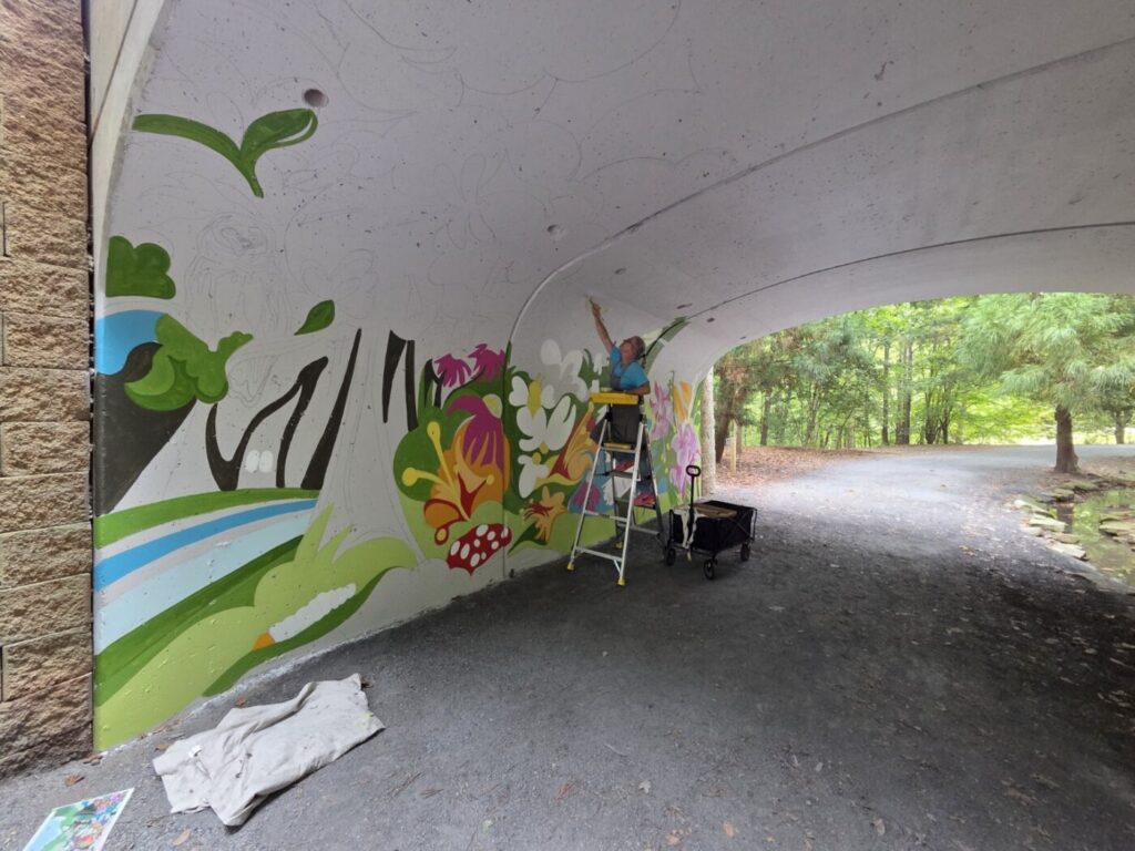 A partially painted mural of flowers and leaves covers one side of the tunnel while an artist works nearby.