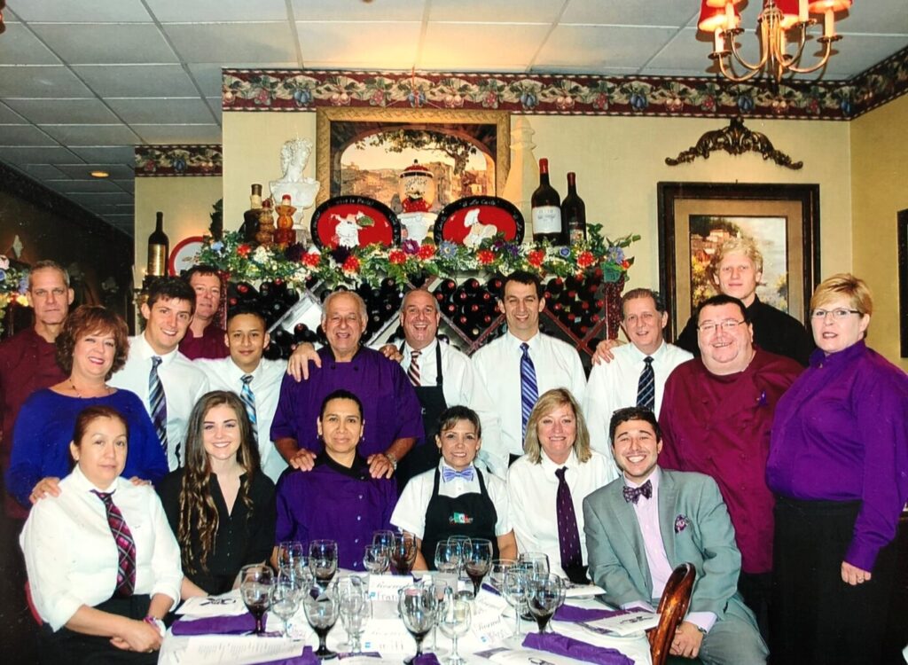 A large group of restaurant staff pose around a table inside a dining room decorated with wine bottles and holiday greenery.